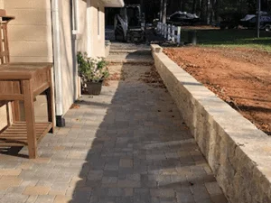 Gray interlocking paver walkway beside a tan segmental stone retaining wall and freshly graded soil
