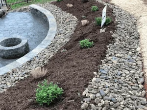 Curved garden bed with dark mulch, river rock border, and young shrubs beside a stone patio edge