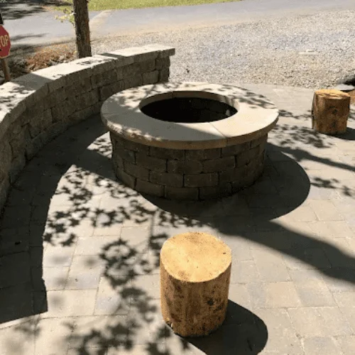 Round stacked stone fire pit with cap stone on gray paver patio, curved block seat wall, and cut log stools