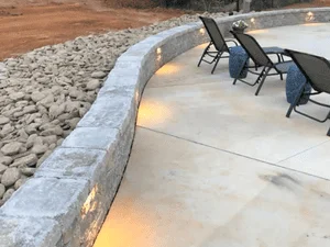 Curved stacked stone retaining wall with warm uplighting beside a concrete patio and river rock bed