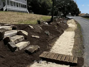 Large natural stone retaining wall with dark topsoil bed, sand pathway, and wood plank steps beside a driveway