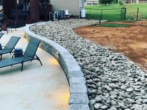 Curved stacked stone retaining wall with capstones beside a concrete patio and gray river rock drainage bed