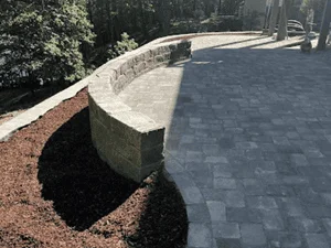 Curved stacked stone retaining wall with cap course beside gray interlocking paver patio and dark mulch bed