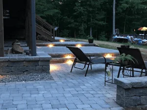 Gray paver patio with stone steps, recessed step lights, and a stacked stone retaining wall at dusk