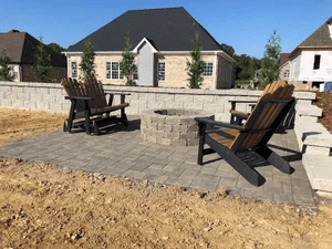 Gray paver patio with stone fire pit, stone seating wall, and dark wooden Adirondack chairs in a newly landscaped backyard