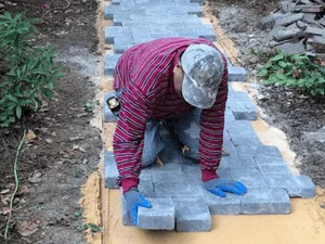 Worker in gloves laying gray interlocking concrete pavers along a curved walkway over yellow-marked sand base