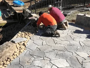 Workers finishing a gray stamped concrete flagstone-pattern patio beside river rock edging and a stone wall