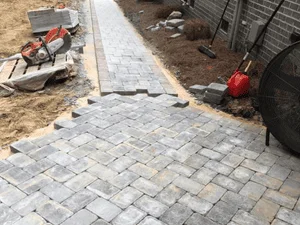 Gray interlocking concrete pavers laid in a herringbone pattern with edge border, beside a red plate compactor and dirt base prep