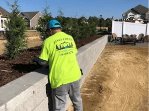 Worker in yellow Piney Creek shirt installing gray block retaining wall beside mulched bed with young shrubs and bare sandy grading