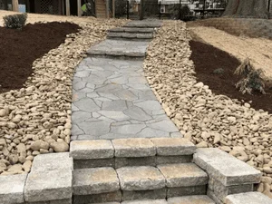 Irregular gray flagstone walkway with tan block steps, stone sidewalls, and river rock border beside mulched beds