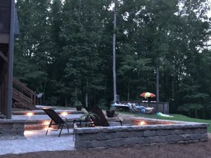 Stone retaining walls with illuminated step and wall lights beside a gray paver patio at dusk, with wooden deck stairs and lounge chairs