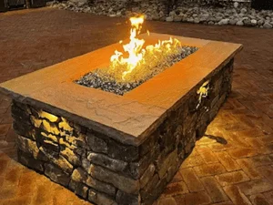 Rectangular gas fire pit with natural stone base, stamped concrete cap, fire glass, and brick paver patio