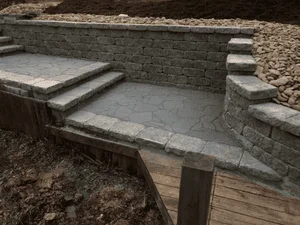 Stacked stone retaining wall with stepped stone landing, stamped concrete patio, and timber edging on a sloped yard
