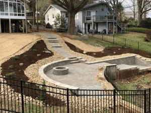 Sloped backyard with curved stone retaining wall, stone fire pit, flagstone steps, gravel, mulch beds, and black metal fence