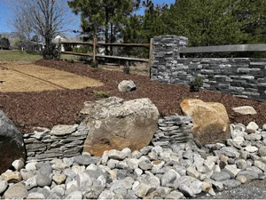 Dry-stacked natural fieldstone retaining wall with large boulder accents, river rock border, and red mulch bed