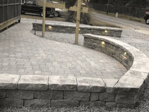 Curved gray paver patio with tiered segmental block retaining walls and recessed wall lights, surrounded by gravel
