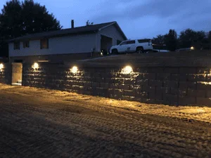 Night view of gray stacked block retaining wall with warm uplights along the base and paver driveway