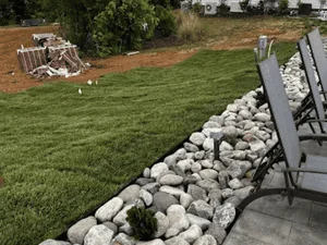 Sod lawn with white and gray river rock border beside gray paver patio and metal lounge chairs